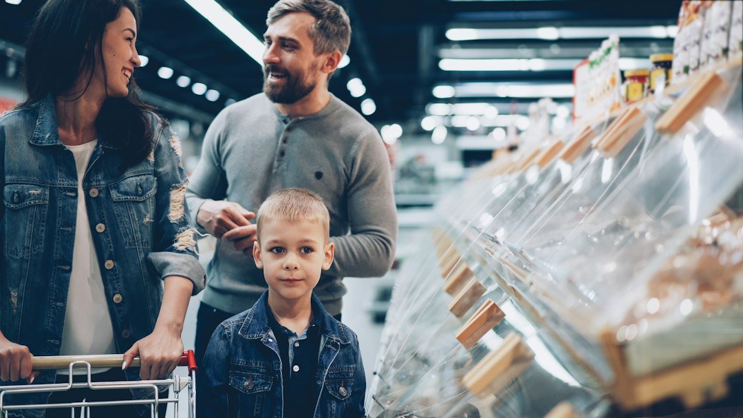 A smiling mother, father, and young child choosing food together in a large supermarket aisle.